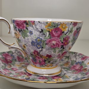 May include: A porcelain teacup and saucer set featuring a vibrant floral pattern with pink, yellow, and blue flowers on a light background. The cup has a curved handle and a gold-trimmed rim, base, and handle edge, matching the gold-trimmed saucer.
