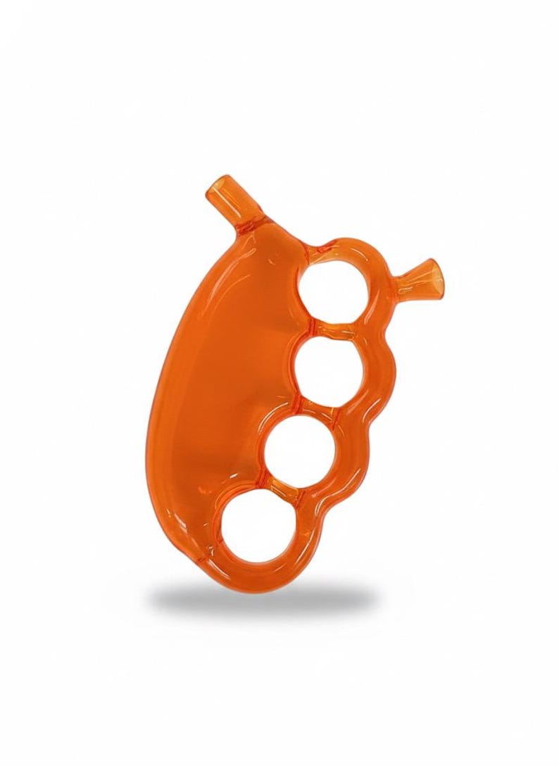 May include: An orange glass pipe with a brass knuckle design. The pipe features four finger holes and two angled mouthpieces. The glass is a translucent orange color, and the overall shape is ergonomic.