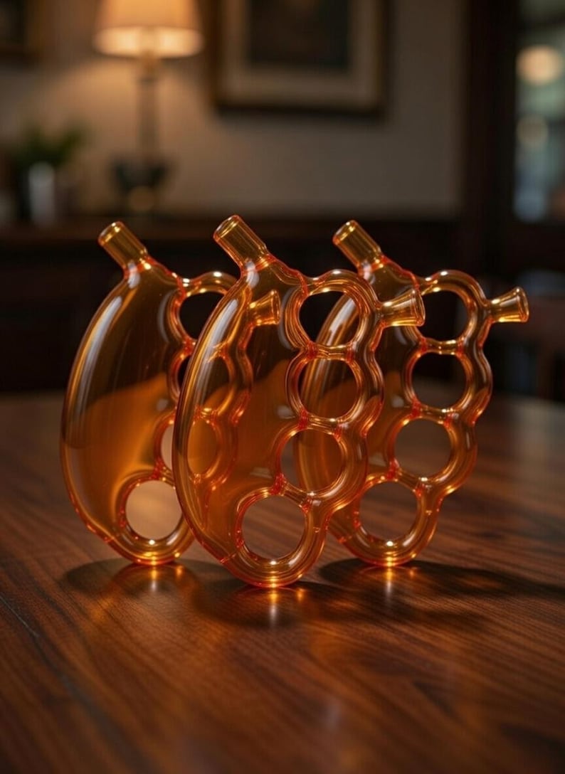May include: Three amber-colored glass decorative objects. Each object has a rounded shape with a series of circular cutouts and a small spout. The objects are arranged on a wooden surface, with soft lighting in the background.