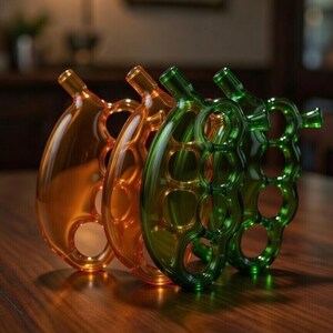May include: Three decorative glass pieces in shades of amber, orange, and green are arranged on a wooden surface. Each piece has a unique, open design with a rounded shape and a small spout.