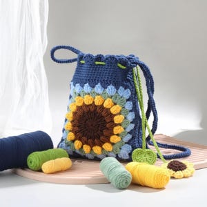May include: A handmade blue crochet drawstring bag with a sunflower design. The sunflower is made with brown, yellow, and green yarn. The bag has a drawstring closure and a green cord. Several spools of yarn in various colors are next to the bag.