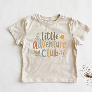 Könnte beinhalten: Beiges kurzärmeliges T-Shirt mit dem bunten Schriftzug „little Adventure Club“. Das Design umfasst eine Sonne, einen Regenbogen und Blätter. Das Shirt ist von Tiny Nest Apparel.