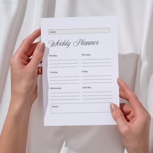 May include: A white weekly planner with the words "Weekly Planner" in elegant script. The planner has sections for each day of the week, a space for notes, and a section to write a name. The planner is held by two hands.