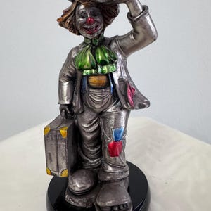 May include: A silver-colored clown figurine with a hat, green necktie, and a suitcase. The clown has a red nose and a painted smile. The figurine is standing on a black base.