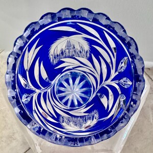 May include: A cobalt blue glass bowl with a scalloped rim. The bowl features intricate white floral and leaf designs. The center has a starburst pattern. The bowl is likely a decorative piece.