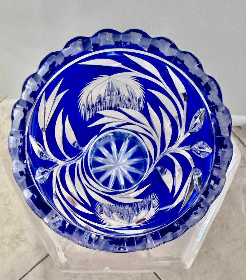 May include: A cobalt blue glass bowl with a scalloped rim. The bowl features intricate white floral and leaf designs. The center has a starburst pattern. The bowl is likely a decorative piece.