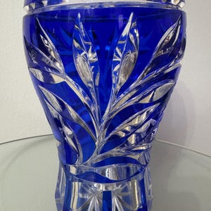 May include: A cobalt blue crystal vase with intricate leaf-like carvings. The vase has a scalloped rim and a faceted base. The design features detailed cuts that reflect light, creating a sparkling effect. A decorative piece for home decor.