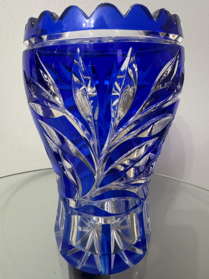 May include: A cobalt blue crystal vase with intricate leaf-like carvings. The vase has a scalloped rim and a faceted base. The design features detailed cuts that reflect light, creating a sparkling effect. A decorative piece for home decor.
