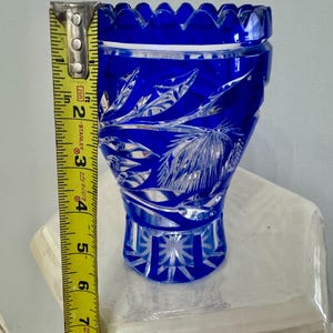 May include: A cobalt blue crystal vase with intricate leaf and geometric patterns. The vase is approximately 6 inches tall, with a scalloped rim. The cut glass design reflects light, creating a sparkling effect. A decorative piece for home decor.