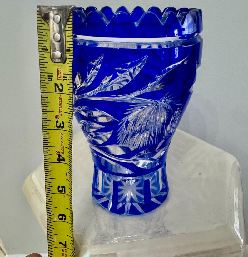 May include: A cobalt blue crystal vase with intricate leaf and geometric patterns. The vase is approximately 6 inches tall, with a scalloped rim. The cut glass design reflects light, creating a sparkling effect. A decorative piece for home decor.