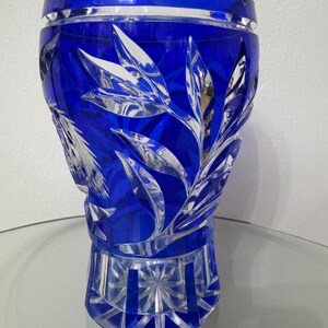 May include: A cobalt blue crystal vase with a leaf design. The vase has a scalloped rim and a geometric base. The leaf design is etched into the crystal, creating a contrast with the smooth, polished surface.