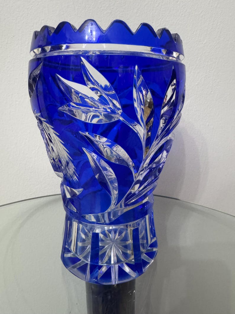 May include: A cobalt blue crystal vase with a leaf design. The vase has a scalloped rim and a geometric base. The leaf design is etched into the crystal, creating a contrast with the smooth, polished surface.