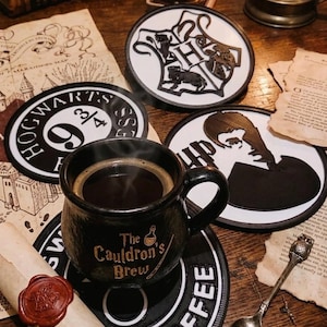 Op de afbeelding: Een zwarte koffiemok met "The Cauldron's Brew" erop staat op een zwarte onderzetter met "COFFEE". Er zijn verschillende Harry Potter-onderzetters te zien, samen met een opgerold perkament en een lepel.