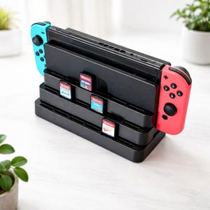 Peut inclure: Station d'accueil de console noire avec une manette bleue et rouge attachée. La station d'accueil comporte des emplacements pour les cartouches de jeu. Les cartouches sont visibles dans les emplacements. L'arrière-plan est une surface de couleur claire.