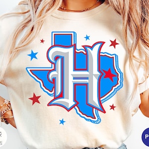Può includere: Una t-shirt color crema con una grande lettera H stilizzata in bianco con ombreggiature grigie e un contorno rosso. Dietro la lettera si trova una sagoma blu dello stato del Texas, accentuata da diverse piccole stelle rosse e blu.