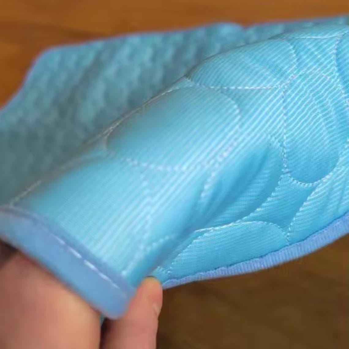 May include: Close-up of a light blue quilted fabric with a diamond pattern. The fabric has a soft, textured appearance and is edged with a matching blue trim. The stitching is visible, creating a subtle design.
