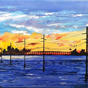 May include: An acrylic painting of a sunset over water. The sky transitions from blue to orange and yellow, with clouds. Silhouetted utility poles and a bridge are in the foreground. The water is a deep blue.