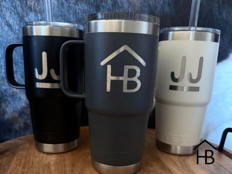 May include: Three insulated tumblers in black, charcoal, and white, each with a silver rim and base. The black tumbler has a handle and the initials "JJ" etched in silver. The charcoal tumbler features a house logo with "HB" in silver. The white tumbler also has "JJ".