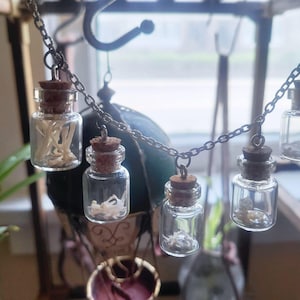 May include: A necklace featuring a silver-toned metal chain with five small, clear glass vials hanging from it. Each vial is sealed with a brown cork stopper and contains small, white, organic bone fragments inside, creating a unique accessory.