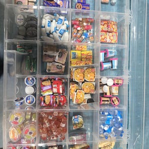May include: A clear plastic organizer filled with miniature food items. The compartments hold tiny boxes of cereal, bottles of milk, snacks, and other grocery products. The items are arranged neatly, showcasing a variety of colors and packaging designs.