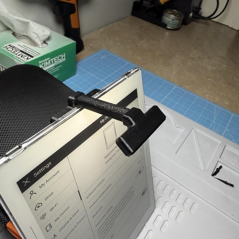 May include: A black tablet holder attached to a tablet, displaying a settings menu. The holder is made of a textured material and is positioned at an angle. A green box of Kimtech tissues is in the background.