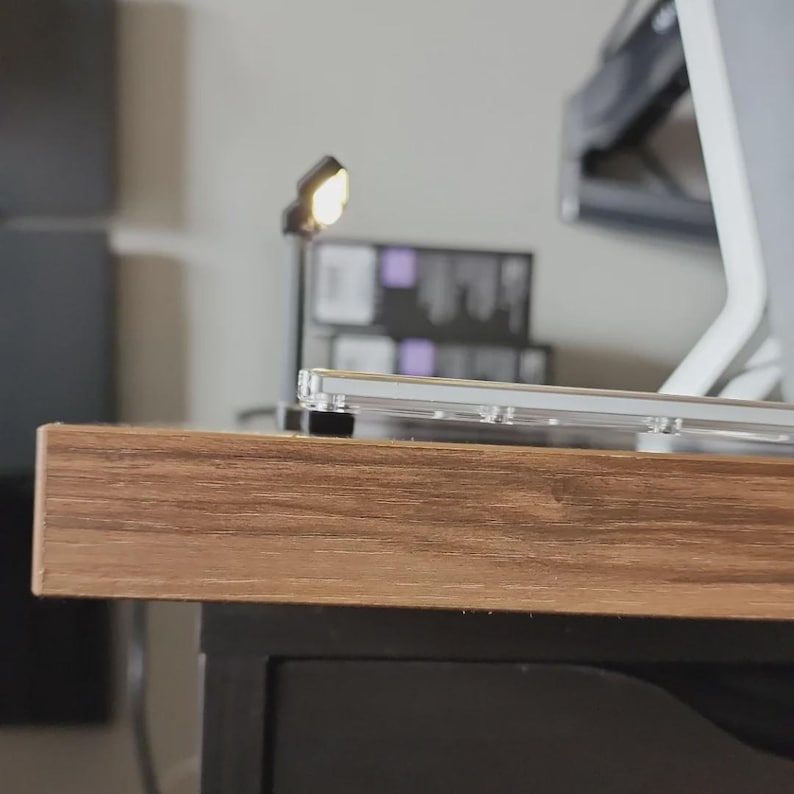 May include: A close-up shot of a wooden desk with a clear, rectangular object on top. A small, adjustable desk lamp is lit, casting a warm glow. The desk has a dark, black cabinet underneath. A computer monitor is visible in the background.