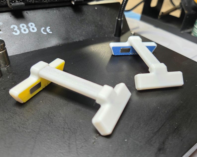 May include: Three white and yellow electronic components with a T-shaped design are arranged on a black surface. The components have a rectangular base with yellow and blue accents. The number "388 ce" is visible in the background.