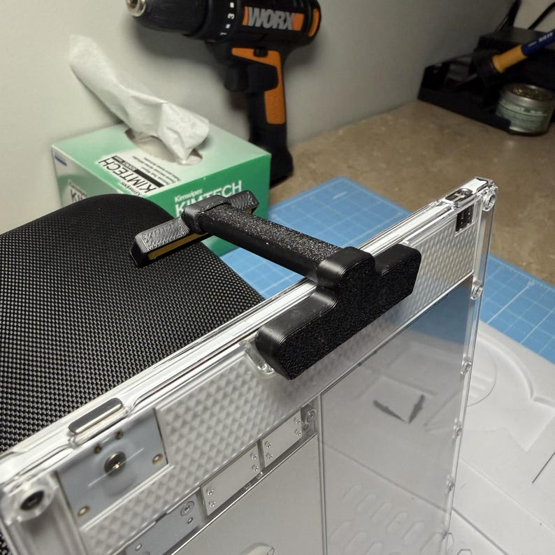 May include: A black and gray tool is attached to a clear plastic case. The tool has a textured handle and two black pads. A drill and a box of tissues are in the background.