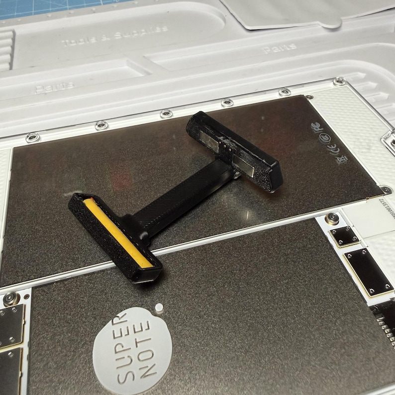 May include: A black and yellow tool with a T-shaped design, resting on a metallic surface. The tool has a textured handle and a yellow stripe on one end. The words "SUPER NOTE" are printed on a white sticker.