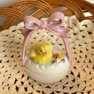 May include: A ceramic Easter egg-shaped trinket box with a yellow chick inside. The box has a pink bow handle and is decorated with small flowers. The egg is white with carved details.