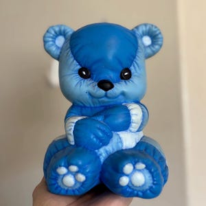May include: A blue ceramic teddy bear figurine with a smiling face, black eyes, and a black nose. The bear has white accents on its arms and legs, and its paws feature white paw prints. The bear is sitting with its arms crossed.