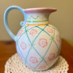 May include: A white ceramic pitcher with a light blue handle and rim. The pitcher is decorated with pink roses and a blue crisscross pattern. It sits on a cream-colored, crocheted doily. The pitcher is approximately 8 inches tall.