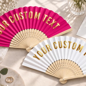 May include: Two folding hand fans with light wood ribs and intricate cut-out details. One fan is vibrant hot pink and the other is white. Both feature the text YOUR CUSTOM TEXT printed in a metallic gold foil font across the pleated fabric surface.