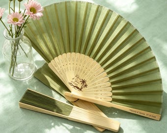 May include: Two folding hand fans with light wood frames and olive green fabric. One fan is open, displaying a palm tree graphic and the text Victoria & Steven 07.29.2028. The other fan is closed, showing the same text on the wooden handle.