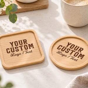 Pode incluir: Dois porta-copos de madeira clara, um quadrado e outro circular, cada um com o texto gravado em castanho escuro "YOUR CUSTOM Logo / Text" centrado na superfície. Os porta-copos têm um acabamento liso e bordas elevadas para estabilidade.