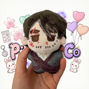 Può includere: Una piccola bambola di peluche con capelli castano scuro, tratti del viso ricamati tra cui una cicatrice sopra l'occhio destro e un abito grigio scuro con un design a cinghie incrociate. La bambola ha una carnagione chiara e una testa rotonda.
