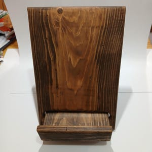 May include: A stained wooden wall-mounted phone holder with a shelf. The wood grain is visible, and the holder is a warm brown color. The holder is rectangular with a small shelf at the bottom.