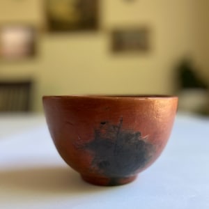 May include: A small, handmade terracotta bowl with a rounded shape. The bowl has a reddish-brown exterior with a dark, mottled pattern near the base. The interior is a darker shade of brown.