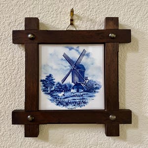 May include: A framed ceramic tile featuring a blue and white windmill scene. The tile is set within a dark brown wooden frame, secured with metal studs at each corner. A small metal hook is attached for hanging.