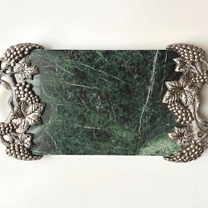 May include: A rectangular serving tray with a green marble surface and ornate silver-colored handles. The handles are decorated with grape clusters and leaves, adding a touch of elegance. The tray is ideal for serving appetizers or displaying decorative items.