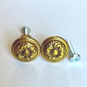 May include: Two antique-style, round brass cabinet knobs with a floral design. The knobs are a warm gold color and have a detailed, embossed pattern. A silver screw is included for installation. Hardware for furniture.