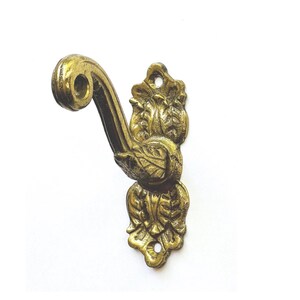 May include: A brass-colored metal wall hook featuring a curved arm and a decorative backplate with embossed leaf patterns. The vertical mounting plate includes two circular screw holes at the top and bottom for secure attachment to a flat surface.