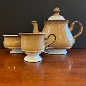 May include: A ceramic tea set featuring a teapot with a lid, a creamer, and a sugar bowl. Each piece has a tan-coloured body with a darker brown gradient near the rim and base, accented by a white pedestal foot and a decorative white band with a pattern.