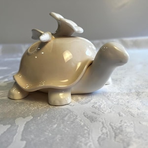 May include: A cream-coloured ceramic turtle figurine with a smooth, glossy finish. The turtle features a stylised flower ornament attached to the top of its shell. The head is extended forward, and the four legs are visible at the base of the shell.