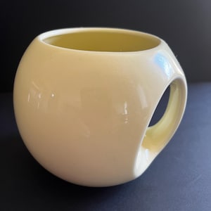 May include: A spherical ceramic mug in a pale cream colour with a smooth, glossy finish. The mug features a unique integrated handle design where the handle is carved directly into the side of the round body, creating a seamless, continuous silhouette.