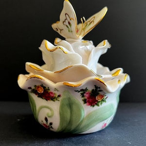 May include: A white porcelain trinket box shaped like a blooming flower with ruffled edges trimmed in gold. The lid features a sculpted butterfly perched atop the petals. The base is decorated with painted green leaves and clusters of red roses.