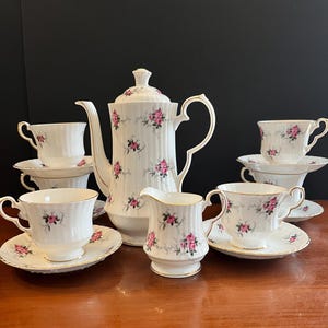 May include: A white porcelain tea set featuring a ribbed texture and a delicate pattern of small pink roses with green leaves. The set includes a tall teapot with a lid, a creamer, and four teacups with matching saucers, all finished with gold trim.