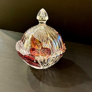 May include: A clear, cut-glass candy dish with a lid. The dish features a ribbed design and a red and orange floral pattern. The lid has a pointed finial. The dish is likely made of crystal.