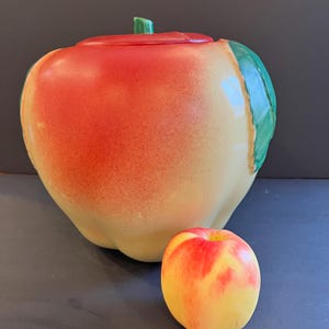 May include: A large, ceramic apple-shaped cookie jar with a red and yellow gradient finish, green stem and leaf accents. A smaller, realistic apple sits beside it. The jar is a decorative kitchen item.