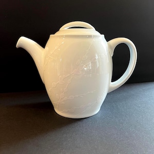 May include: A white porcelain teapot featuring a subtle, embossed pussy willow branch pattern across its surface. The teapot includes a matching lid with a curved handle, a curved spout, and a rounded handle on the side for pouring tea.